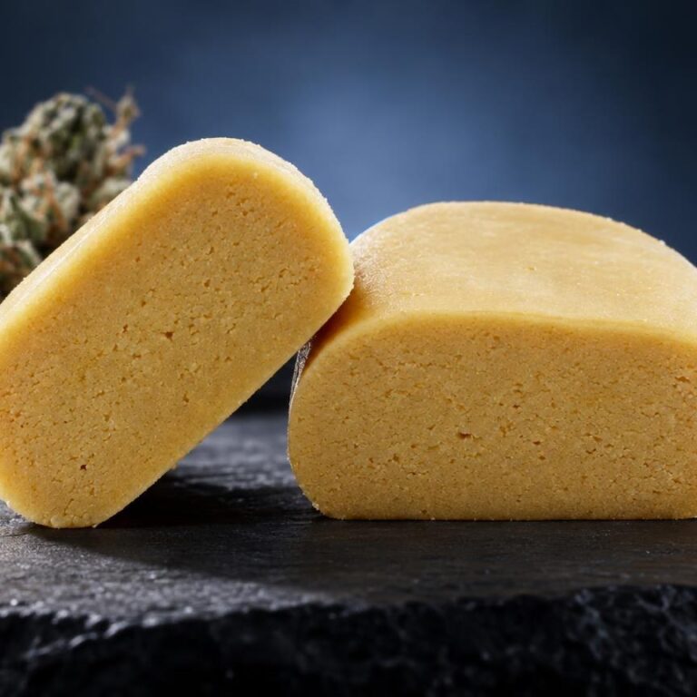 Piatella : deux plaques de hash dorées à texture granuleuse posées sur ardoise noire, avec fleur séchée en arrière-plan